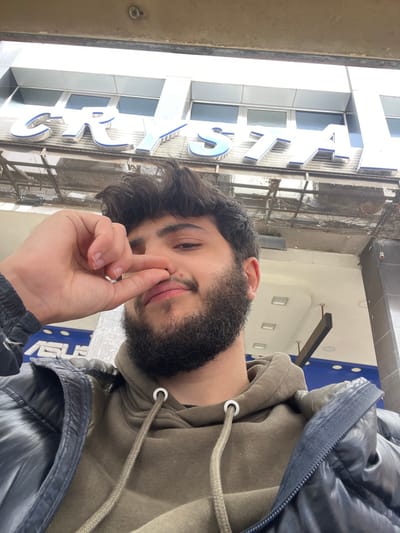A male individual with dark, curly hair and a full dark beard is captured in a medium-close-up, upward-angled shot. He wears a dark, possibly navy, padded jacket over an olive-green hooded sweatshirt with visible white-ringed drawstrings. His right hand is positioned with the thumb and index finger pinching the tip of his nose, while he looks towards the camera with a subtle smile. The background reveals a building facade in Sarouja Municipality, Syria, featuring a large sign with raised, blue-outlined white letters spelling "CRYSTA" (likely part of "CRYSTAL"). Below this, a smaller sign partially displays the white letters "ASUS" on a dark background. A metal grate structure is visible between the two signs.