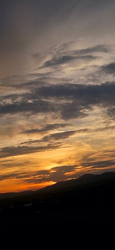A photograph captures a dramatic sunset over a mountainous landscape. The sky is a canvas of swirling clouds, transitioning from deep grays and purples at the top to vibrant oranges and yellows near the horizon. Two faint white contrails from airplanes are visible crisscrossing the sky. Below the sky, silhouetted against the sunset, are the dark, rolling forms of mountains. The foreground is a dark, indistinct expanse, likely representing a valley or plain dotted with subtle hints of structures. The overall impression is one of natural beauty and atmospheric grandeur. The location is identified as Momin Prohod, Bulgaria.