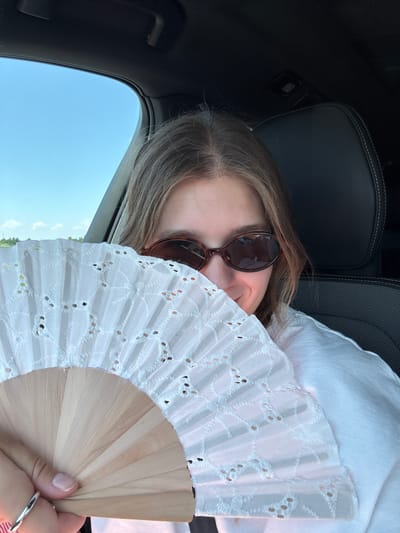 A young woman sits in the passenger seat of a vehicle, holding a white fabric fan with a wooden handle. She is wearing sunglasses and a white shirt. The fan obscures the lower half of her face. The interior of the car, including a black leather headrest, is visible, as is the blue sky and some distant greenery through the window. The scene appears to be taking place outside the city of Zagreb, Croatia, likely on a sunny day.