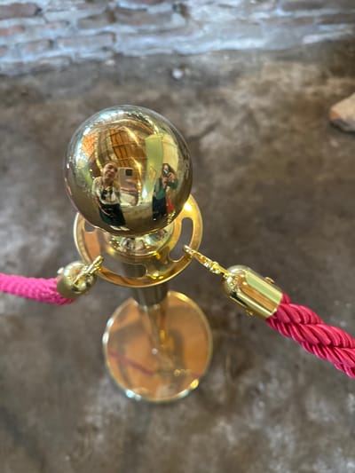 The image shows a close-up, high-angle view of a stanchion with a golden ball topper. The ball reflects two individuals taking selfies. One individual is wearing a white top and a dark skirt, and the other is wearing a green top. The stanchion is part of a velvet rope barrier, with a pink rope visible attached to the stanchion. The background consists of a textured brick wall and a grey concrete floor. The scene appears to be an indoor public space. There is no information or visual evidence in the image to suggest the location is the city of Sremska Mitrovica, Serbia.