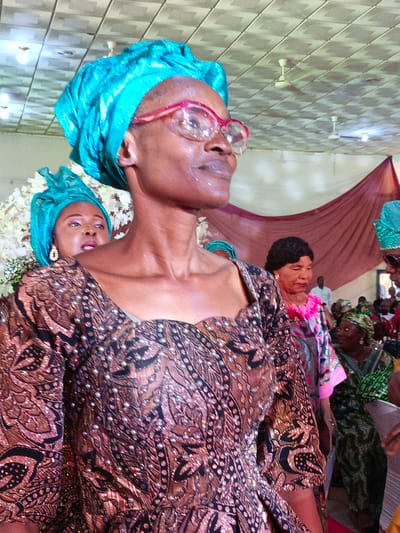 In an indoor setting, possibly in Fagge D2, Nigeria, the image captures several women dressed in vibrant, patterned clothing and headwraps. The main subject is a woman wearing a brown and black patterned dress adorned with small reflective embellishments. She has a bright blue headwrap and wears pink-framed glasses. Another woman in a matching headwrap is visible behind her, partially obscured, wearing earrings. In the background, other women are seen, including one with a floral necklace over a grey and pink patterned dress. The environment includes a tiled ceiling with ceiling fans and what appears to be a draped maroon curtain or backdrop. The overall impression is of a gathering or celebration.