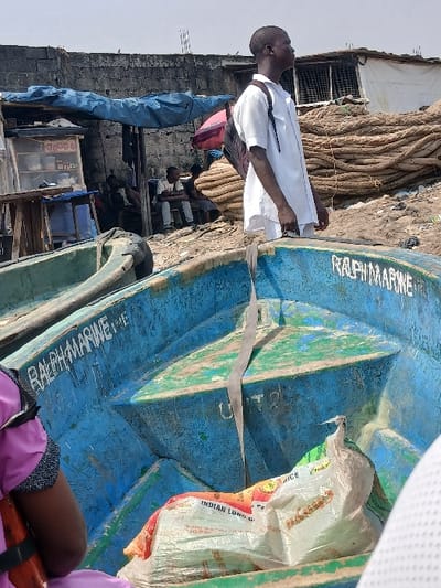 This image depicts a bustling waterfront scene in Alaiyabiagba, Nigeria, characterized by informal market activity and maritime elements under bright, hazy daylight.
In the foreground, the interior of a weathered, blue and green painted boat, likely wooden or fiberglass, occupies the lower central and right portions of the frame. The words "RALPH MARINE" are stenciled in white capital letters on its interior side. Several white sacks, one prominently displaying "INDIAN LONG GRAIN RICE," are placed as cargo within this boat. A grey strap is visible extending vertically from the boat's side down to its floor. Partially visible on the far left, a person wearing a purple garment and an orange life vest strap is seated in an adjacent, obscured vessel.
In the midground, a dark-skinned male, appearing to be a young adult, stands facing towards the upper right of the frame, his gaze directed upwards and away from the camera. He wears a short-sleeved white collared shirt, dark trousers, and a dark backpack on his left shoulder. He appears to be standing in or near a boat, or on the immediate shoreline. Behind him, on the cluttered ground, a makeshift market stall with a blue tarp roof is visible. The stall displays small packaged goods on shelves. Several individuals are seated in this area, some under the blue tarp, others further right under what appears to be a red umbrella or tarp. A substantial pile of thick, coiled ropes, possibly for fishing or mooring, is stacked on the ground to the right of the market stall.
The background consists of a grey concrete block wall, indicative of a permanent structure or boundary. Above and behind this wall, a few rudimentary structures with corrugated metal roofing are visible under a bright, overcast, or hazy sky. The immediate ground appears uneven, sandy, and scattered with debris.