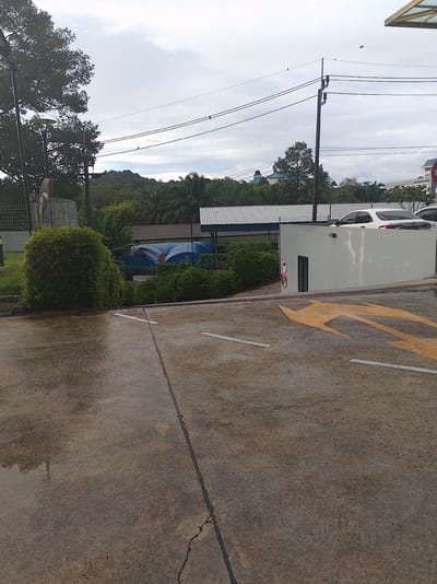 This image shows a wet concrete surface, possibly a parking lot or road, with visible water puddles reflecting trees. In the background, there are several cars, including a white sedan, parked near a building. The scene is set against a backdrop of lush green trees and hills under an overcast sky. Electrical wires and poles are also visible. A yellow arrow is painted on the concrete, indicating a direction. There are no people visible in the image. The presence of a white sedan and what appears to be a public building with a white wall and a blue awning suggest an urban or suburban setting. The style of the building and the surrounding vegetation are consistent with the tropical climate of Phuket City Municipality, Thailand.