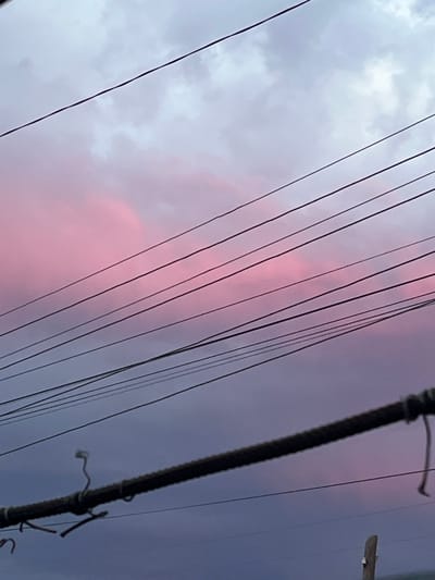 The image depicts a sky with pink and purple clouds, suggesting it was taken during either sunrise or sunset. Several dark utility wires are strung across the frame, obscuring parts of the sky. In the foreground, a thick, dark wire or cable is prominently visible, with a makeshift wire hanger attached to it. A portion of a wooden post is visible at the bottom right corner, also appearing to be part of the utility infrastructure. The scene is suggestive of an urban or semi-urban environment.