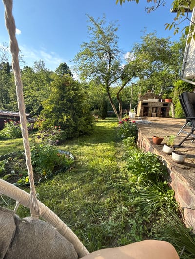 The image presents a backyard scene. It's a sunny day with a clear blue sky visible through the trees. The perspective is from within a hanging chair, the rope and seat are visible in the foreground. The backyard is overgrown with grass and vegetation. There is a stone patio in the background with a brick fireplace or outdoor oven structure. On the patio, there's a chair, some flower pots, and various items stored within the fireplace structure. In the garden, there are various plants, some with red and purple flowers, bordered with a black plastic garden edge. The location is possibly in Ravnishte, Bulgaria, given the prompt's information.