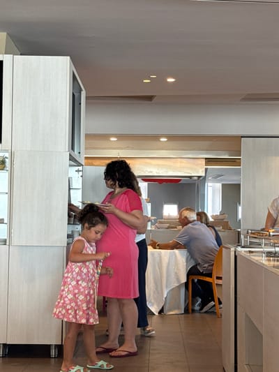 The image depicts an indoor scene that appears to be a buffet or dining area, likely within a hotel or restaurant in Isola delle Femmine, Italy. In the foreground, a young girl in a pink floral dress and light blue sandals is standing next to a woman wearing a bright pink dress. The woman is holding a white plate with some food on it. To the right, a man in a blue polo shirt and dark pants is seated at a table covered with a white tablecloth, with an orange chair beside him. Further in the background, other patrons can be seen, including a woman and a man sitting at another table. There are buffet serving stations visible with various food items and dishes. The overall environment suggests a casual dining setting with clean, modern architecture.