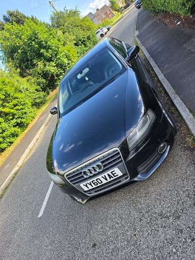 A black Audi A5 S Line, registered YY60 VAE, is parked on a residential street in Shotton, United Kingdom. The car is positioned at an angle, with its front facing slightly towards the right of the frame and its passenger side closest to the camera. The vehicle is parked on the left side of the road, partially overlapping a white painted line that indicates a parking bay. The road surface is asphalt, with some patches of gravel. To the left of the road, there is a grass verge with a concrete curb. Beyond the verge, there are dense green trees and bushes. To the right of the road, there is a paved footpath, a concrete curb, and a hedgerow. In the background, a residential building with a red brick facade and a pitched roof is visible. The sky is blue with some scattered white clouds. The image appears to have been taken on a sunny day.