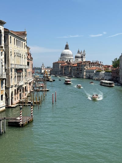 This media file depicts a sunny day in Venice, Italy, likely late morning or early afternoon given the bright light and clear blue sky. The scene is a panoramic view of a canal lined with historic buildings. The architecture is characteristic of Venice, with ornate facades, arched windows, and terracotta tiled roofs. Prominently featured is a large domed building, likely a church or basilica, with smaller domes and spires.
Several boats are navigating the canal, including a larger passenger boat and smaller motorboats. One motorboat is creating a noticeable wake, indicating movement. The canal itself is a murky green, a common color for Venetian waterways, and is lined with wooden posts, some of which are striped with red and white. These posts likely serve as moorings or markers.
The perspective suggests the photo was taken from a higher vantage point, possibly another building or bridge, looking down onto the canal and the surrounding architecture. The overall atmosphere is one of a bustling urban waterway in a historic European city. There is no visible text within the image.