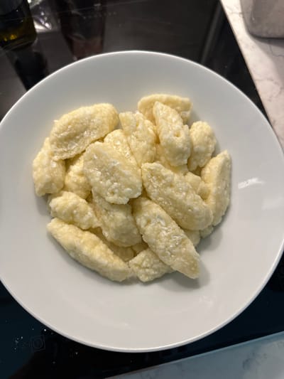 A close-up, eye-level shot captures a white ceramic bowl filled with homemade cottage cheese dumplings, known as "halušky" or "pirohy," on a black stovetop. The dumplings are elongated, irregularly shaped, and a pale yellow color, with some appearing slightly clumped together. The bowl is positioned slightly off-center, taking up most of the frame, and rests on a smooth, reflective black surface that appears to be an induction cooktop. In the blurred background, a sliver of a marble countertop is visible to the right, and to the left, dark reflections suggest kitchen appliances. The scene is well-lit, highlighting the texture of the dumplings. This image was likely taken in a residential kitchen, possibly in Helsinki, Finland, given the context provided, though the specific location cannot be definitively determined from the visual information alone.
