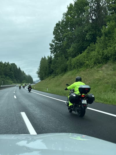 A group of motorcyclists is traveling in formation on a wet highway on an overcast day. The perspective is from a vehicle following the motorcycles. The lead motorcycle in the foreground has a rider wearing a bright yellow jacket and helmet, with black saddlebags and a black top case. The motorcycle's license plate reads "KNS 33S2". Several other motorcycles, also with riders in yellow gear, are visible further ahead on the highway. The highway is flanked by a steep, grassy embankment on the right, covered in lush green trees and foliage. The road itself is dark asphalt with white lane markings. The sky is a uniform light gray, indicating cloudy weather. The overall scene suggests a motorcycle tour or ride in a rural, mountainous area.