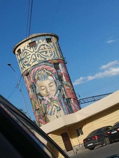 The image shows a tall, cylindrical water tower in the city of Shymkent, Kazakhstan, adorned with a large, colorful mural. The mural depicts a woman's face, wearing a traditional headdress, holding a bouquet of pink flowers. The top section of the tower features geometric patterns in blue and yellow. The tower is tilted, suggesting a unique architectural style or an optical illusion. In the foreground, the reflection of a vehicle's interior is visible on a dark, shiny surface, likely a car window or body panel. To the right of the tower, a light yellow building with a corrugated roof runs horizontally across the frame. Two black sedans are parked in front of this building. The sky is clear blue with a few scattered clouds. A streetlamp and power lines are visible near the tower. A metal bridge-like structure is seen in the background, partially obscured by the yellow building. The overall scene appears to be a street view in an urban environment under a bright sky.