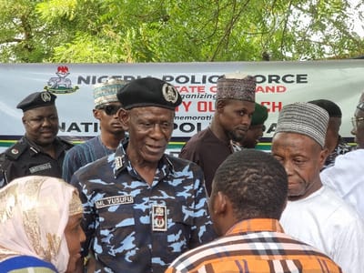 The image shows a group of people standing in front of a banner that reads "NIGERIA POLICE FORCE STATE COMMAND ORGANIZING MY OWN CH MEMORATIO POLI." The banner is located in Maiduguri, Borno State, Nigeria. The group is composed of mostly men, some are wearing traditional Nigerian clothing and headwear, including a few in police uniform. In the center of the group is a man in a police uniform and cap, speaking to the people. The man is identified by a nametag as "ML YUSUFU". The individuals are interacting in a gathering setting, possibly a community event or gathering. There are trees and greenery in the background.