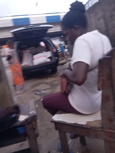 In Port Harcourt, Nigeria, a blurred image captures a street scene. In the foreground, a woman in a white t-shirt and burgundy leggings sits on a wooden chair. She holds a pink purse in her lap. To her left, another woman in an orange and grey patterned dress stands near the open trunk of a dark-colored SUV, which is loaded with sacks. In the background, a person in a blue shirt walks away from the vehicle. The environment appears to be an outdoor market or roadside area, with concrete ground and hints of buildings and a striped overpass.