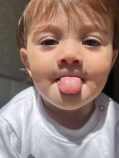 A close-up image shows a baby with fair skin, light brown hair, and brown eyes. The baby is sticking their tongue out. The baby is wearing a white garment with a visible snap button. The background appears to be a neutral gray color. No definitive details indicate the location is in Elena, Bulgaria.