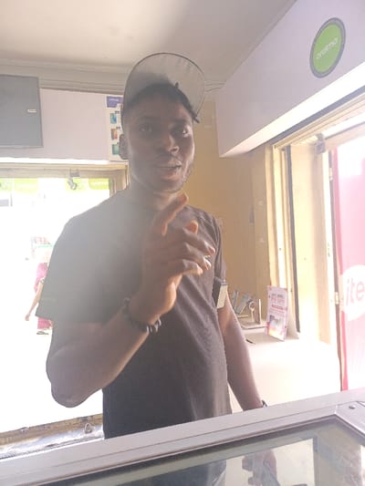 The image captures an indoor commercial setting, likely a retail establishment, located in Fagge A, Nigeria. The primary subject is a male individual, appearing to be of African descent, positioned from the chest up and looking directly at the camera. He wears a dark short-sleeved t-shirt, a light-colored baseball cap worn backward, and a dark beaded bracelet on his left wrist. His right hand is raised with the index finger extended, pointing slightly upward.
The background displays various commercial elements. To the left, a dark rectangular unit is mounted high on a white wall. Below it, vertical posters for "VIVO" mobile phones are visible on a white panel. An "oratmo" logo sticker is affixed to a door frame. A brightly lit doorway or opening on the left reveals a blurred, partially visible figure in a pink garment, suggesting an outdoor or brightly lit area beyond. To the right of the subject, a white horizontal panel, possibly an air conditioning unit or a sign, bears a circular green "oraimo" logo. Below this, a red and white promotional poster is displayed, containing partially visible text such as "LIVE MORE, PAY LESS" and "ite". The wall in the background appears light brown/beige. The foreground shows the top edge of a counter or display case. The scene is brightly lit, with some areas of overexposure.