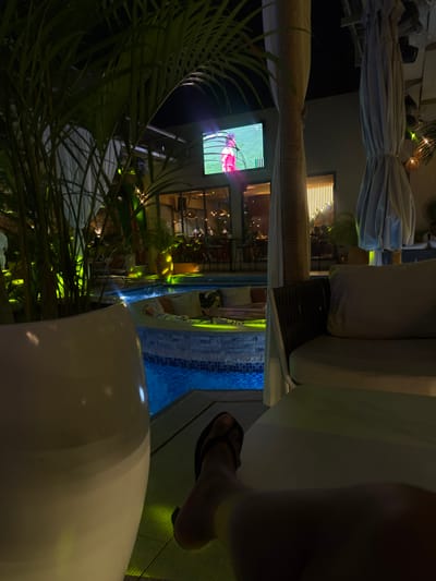 A nighttime view from an outdoor patio, likely near a pool or water feature given the tile design and reflection, includes a person's leg and foot in a dark flip-flop in the foreground on the right side. The patio is furnished with a large white cushion sofa and a white, oversized plant container. In the distance, across a pool area, a TV screen is visible, displaying an image that seems to show a sports game or broadcast. The scene is heavily obscured by plant foliage on the left, which gives a sense of privacy or seclusion. The location is purportedly in Itirin, Nigeria, but nothing is present in the image that substantiates this claim.