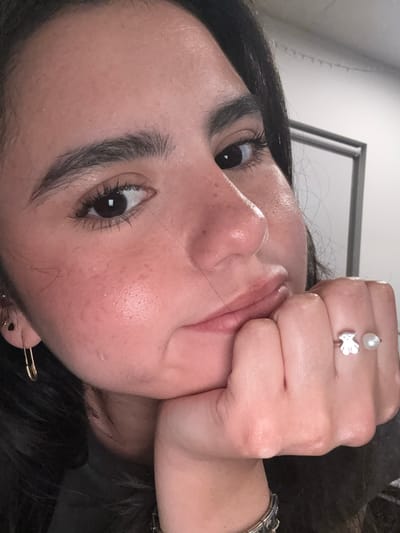 A close-up photograph depicts a person's face with dark hair. The person's face is tilted towards the right, and their hand is positioned against their cheek. The person's skin tone appears fair. The person is wearing a ring with a pearl and a fish charm. The setting is in the United States of America. There is no further information in the image to determine the location in Austin, United States of America.