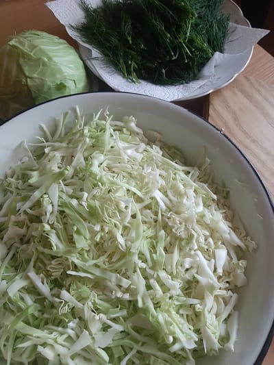 This image shows a close-up view of freshly prepared ingredients for a meal, likely a salad or soup, laid out on a wooden table. The primary subjects are food items: a large bowl filled with thinly shredded white cabbage, a smaller bowl containing a bunch of fresh green dill resting on a white paper towel, and a whole head of green cabbage placed on the table. The shredded cabbage fills most of the frame, appearing crisp and vibrant with light green and white tones. The dill is dark green and feathery, with some of its stems visible. The whole cabbage head is a lighter shade of green and has visible leaf layers.
The scene is set indoors, on a wooden surface that appears to be a table. The lighting suggests it is daytime, possibly natural light filtering in, as there are no harsh shadows. The overall impression is one of food preparation, with the ingredients ready to be combined. There are no people visible in the image. The composition is focused on the textures and colors of the vegetables.
There is no visible text in the image. The setting, Lviv, Ukraine, is provided as context but is not visually represented by any specific landmarks or cultural cues within the image itself. The image conveys a sense of freshness and the beginning of a cooking process.