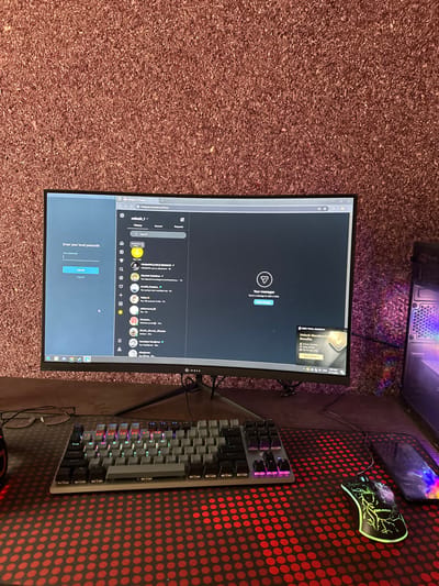 The image shows a computer setup with a monitor displaying a screen with a message stating "Your messages: Send a message to start a chat." Various icons and a log-in prompt are also visible. A keyboard with RGB lighting is positioned to the right of the monitor, sitting on a red and black patterned mousepad. A gaming mouse is on the upper right and a PC case is slightly visible above the monitor. Glasses are visible near the keyboard. The entire setup is placed on a brown carpet.