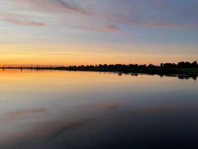 A serene, wide river reflects the soft hues of a twilight sky over Sovetsk, Russia. A long, metal truss bridge spans the river in the distance, its structure silhouetted against the fading orange and pink glow. The opposite bank is lined with a dense band of dark trees and scattered buildings, their shapes softened by the low light. The water's surface is remarkably still, mirroring the sky and the distant landscape with perfect clarity, creating a sense of peaceful symmetry. The sky transitions from a warm orange near the horizon to cooler blues and purples overhead, with wispy clouds catching the last light of the day. The overall impression is one of tranquility and natural beauty at the close of day in this Russian city.