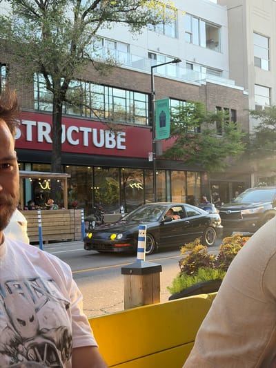 The image depicts a street scene in Montreal, Canada, possibly on Rue Saint-Denis, with a focus on a restaurant or storefront with the prominent red signage reading "STRUCTUBE". The sun is setting, casting a warm glow on the scene. In the foreground, a person is partially visible, wearing a white t-shirt with a graphic print. To the left, an outdoor seating area with wooden dividers and blue and white bollards is set up. A black coupe with distinctive yellow headlights is driving on the street in front of the establishment. Behind the coupe, a darker SUV is also visible. Trees line the street, and modern buildings with large windows form the backdrop. A green banner with text and an icon is hanging from a lamppost. The overall atmosphere suggests a lively urban environment during the evening.