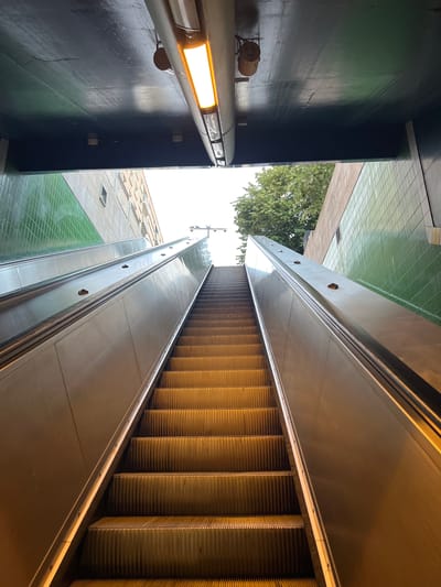 The image shows an upward view of a wide, modern escalator leading out of an underground passage towards an open, bright sky and trees. The escalator steps are made of metal with ribbed textures. On either side of the escalator, there are polished metal walls and a green tiled wall section. Above the escalator entrance, a dark, covered ceiling with a fluorescent light fixture is visible. The surrounding environment suggests an urban setting, possibly a public transportation station. The overall impression is one of movement and transition. The location is not determinable from the image alone.