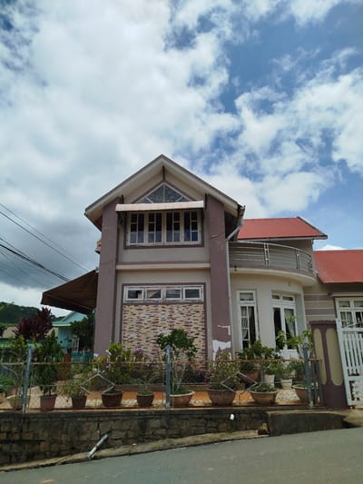 This is a full shot, eye-level, exterior photograph of a two-story house with a red-tiled roof, located in the city of Xuân Sơn, Vietnam. The house has a light brown and white facade with brown trim around the windows and doors. The front of the house features several windows and a balcony on the second floor, with a curved metal railing. The ground floor has a large, arched doorway with white double doors and is lined with potted plants. A white metal gate is visible on the right side of the house. The house is surrounded by a fence with a stone wall at its base, and more potted plants are arranged along the fence. The sky is blue with scattered white clouds, and a green hill is visible in the background. The street in front of the house is paved with asphalt.