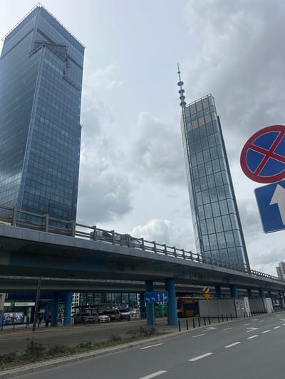 The image shows a street scene in Warsaw, Poland, featuring modern architecture and infrastructure. Two prominent skyscrapers dominate the skyline. The skyscraper on the left is a dark glass-clad building. The skyscraper on the right is taller and also glass-clad, with a distinctive antenna array at its peak and a section of its upper facade appearing to be tilted or canted.

A multi-level overpass or elevated roadway runs horizontally across the middle of the frame, supported by blue pillars. The overpass has a wooden railing along its edge. Below the overpass, a street level is visible with several cars parked. A bus is also visible. On the right side of the image, there are two traffic signs: one circular with a red border and a blue interior showing a red "X" (indicating no stopping or parking), and a blue rectangular sign with a white arrow pointing upwards.

The sky is overcast with clouds, suggesting a neutral or possibly cool weather day. The environment appears to be an urban setting, with evidence of ongoing development or maintenance, suggested by the wooden railing on the overpass. One of the lower buildings visible beneath the overpass has a sign that reads "BNP PARIBAS". The overall impression is one of a bustling, modern city with significant infrastructure.
