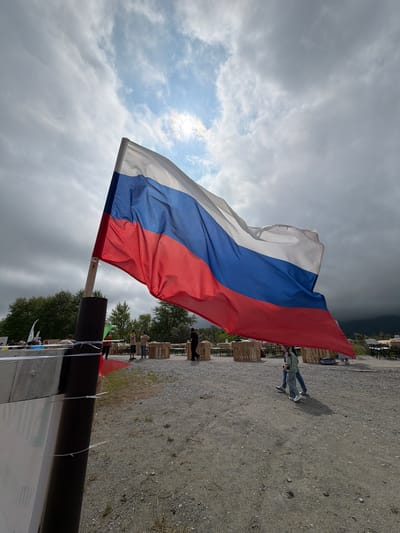 An outdoor scene features a large Russian flag prominently displayed, waving in the wind against a cloudy sky with patches of sunlight. The flag, composed of white, blue, and red horizontal stripes, is attached to a wooden pole wrapped in dark material. In the background, several people are visible, some standing near wooden crates and tables, suggesting an event or gathering. Trees and hills form the distant landscape. The ground is covered with gravel and dirt, and some white tape barriers are present in the foreground. The scene appears to be in an open, possibly rural or park-like setting, with a few structures and seating areas visible. The overall impression is one of a public outdoor event under a dynamic sky.