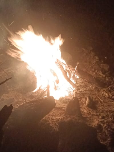 A vibrant campfire burns intensely in the darkness, illuminating its immediate surroundings. Branches fuel the flames, casting a warm glow on the ground. Two dark shapes, likely logs, are in the foreground, partially obscured by the low light. The ground appears to be dirt or sand, dotted with small rocks. The scene suggests a camping or outdoor setting during the night.