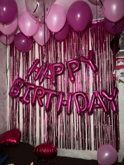 This image depicts a birthday party setup. A metallic fringe curtain in a bright pink or fuchsia color serves as a backdrop. In front of the curtain, large foil balloons spell out "HAPPY BIRTHDAY" in a matching pink color. Several round balloons in shades of pink and fuchsia are scattered around the scene, some hanging from the ceiling and others on the floor. Two heart-shaped balloons, also in pink, are visible at the bottom left of the image, resting on what appears to be furniture. The overall impression is a festive and colorful celebration space. The image does not contain any identifiable landmarks or clues that would indicate its specific location within the city of Samara, Russia.