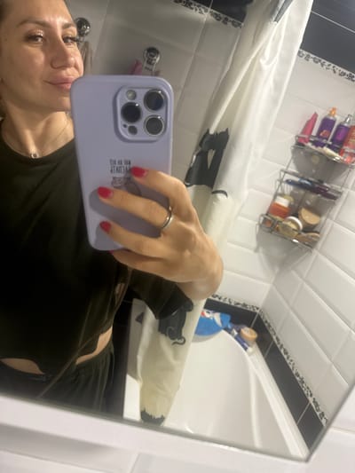 The media file shows a woman taking a selfie in a bathroom. The woman is in the foreground, holding a purple phone with a camera bump and a silver ring on her finger. She is wearing a dark green top and a necklace. Her makeup is visible, including mascara and lipstick. In the background, a shower curtain with a black and white design is visible, along with a bathtub and tiled walls. A shelf with toiletries, including bottles of shampoo and other products, is mounted on the wall. There is also a blue bottle with a cartoon character on it, possibly a cleaning product. The overall setting appears to be a home bathroom. The image is taken from a low angle, with the camera tilted upwards. The lighting suggests it is daytime. There is some text on the phone case, which is difficult to read due to the angle and resolution.
