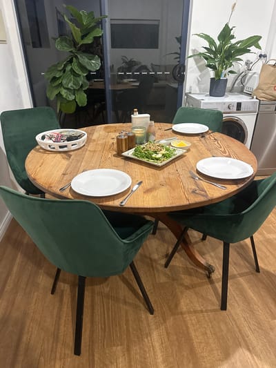 The image depicts a dining room scene in Bonville, Australia, featuring a circular wooden dining table set for at least four people. The table is adorned with white plates, silverware, a serving dish of salad, and decorative items including salt and pepper shakers and a candle. Four green velvet dining chairs with black legs surround the table. In the background, through a glass door, a glimpse of a living room area can be seen. To the right of the dining area, a washing machine and dryer are visible, along with a large potted peace lily plant. A large fiddle leaf fig tree is situated to the left, near the glass door. The flooring is light-colored wood laminate.