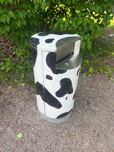 A cow-patterned trash receptacle stands on a gravel path in front of a dense green bush. The receptacle is cylindrical and painted white with irregular black splotches, mimicking the coat of a Holstein cow. It has a silver metal top with a slot for depositing waste. The bush behind it is lush and leafy, with sunlight filtering through its branches. The ground is covered in small, varied-colored gravel. The scene appears to be outdoors, possibly in a park or along a walking trail, in Helsinki, Finland. There are no people or animals visible in the image.