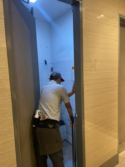In North Jakarta, Indonesia, a male cleaner, wearing a light blue polo shirt, dark apron, and a baseball cap, is seen pushing open a door to a small, tiled room. The room appears to be a utility closet or a small restroom, with a mop and broom leaning against the wall. The cleaner has a bottle of disinfectant clipped to his belt. The exterior walls of the room and the surrounding hallway are lined with beige, wood-grain textured panels. The lighting in the room is stark and blue-toned, contrasting with the warmer lighting of the hallway.