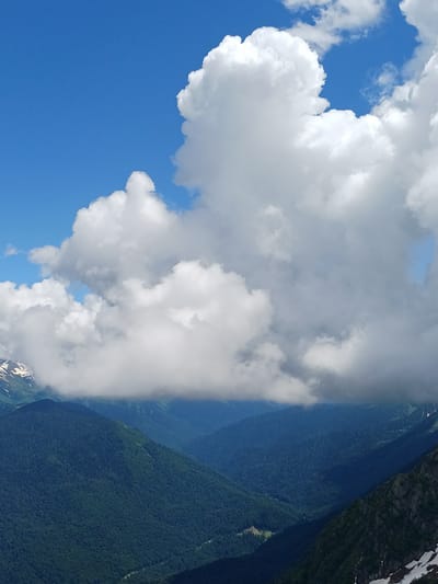 This image captures a scenic view of a mountainous region, likely near Estosadok, Russia, under a bright blue sky dotted with dramatic white clouds. The foreground features the jagged slopes of a dark, rocky mountain covered in patches of snow, contrasting with the deep green, densely forested slopes of lower mountains in the mid-ground. These forested mountains recede into the distance, forming a series of valleys shrouded in a soft, atmospheric haze. A thick band of clouds hovers low over the mid-ground mountains, partially obscuring the view and creating a sense of depth. The sky above is a vibrant blue, with large, fluffy cumulus clouds dominating the upper portion of the frame, their bright white surfaces catching the sunlight. No people or man-made structures are clearly visible, emphasizing the natural grandeur of the landscape.