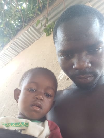 A close-up image taken in Lafia, Nigeria, features an adult male and a young child. The adult male, positioned on the right, is bare-chested, has dark skin, short dark hair, and facial stubble. He looks directly at the camera with a neutral expression. To his left, a dark-skinned toddler with short dark hair and a small silver-colored earring in their left ear is partially visible, also looking towards the camera with a slightly open mouth. The child wears a multi-colored garment featuring white, green, and red sections. The background consists of a light-colored, textured wall. Above the wall, a section of corrugated metal roofing is visible, partially obscured by green foliage. The scene is illuminated by natural light.