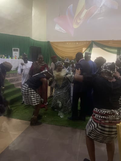 A group of people are gathered indoors, possibly for a celebration or event, in Markurdi, Nigeria. The scene is dimly lit, with a projection screen displaying a colorful abstract design on the wall above. The room features green and yellow drapery adorning the walls, suggesting a festive atmosphere. A few individuals are actively dancing or moving, their attire reflecting a mix of traditional and modern styles. Some are wearing traditional Nigerian clothing, including vibrant patterned fabrics and headwraps, while others are in more formal attire like suits. One person in the foreground is wearing a black top and a black and white checkered skirt with beaded accents around the waist, engaged in a dance. Another individual in a white robe and traditional cap is also visible. The flooring appears to be a combination of tiled and carpeted areas, with a faux grass mat placed in the central area where some of the dancing is taking place. The presence of a white air conditioning unit and what appears to be a table with bottles and glasses in the background suggests this is an indoor venue.