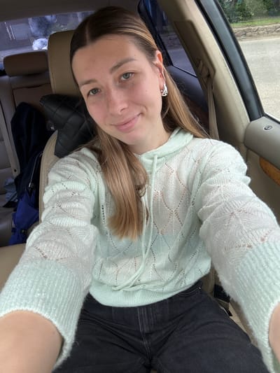 A young woman with light brown hair and freckles is taking a selfie in the passenger seat of a car. She is wearing a pale green hooded sweater with a diamond pattern and black jeans. Her left arm is resting on her lap, and her right arm is extended to take the photo. The interior of the car is visible, including the beige leather seats and a portion of the dashboard. Outside the car window, greenery and a paved road can be seen. The image appears to have been taken during the daytime.