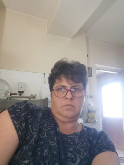 The image shows a woman wearing a dark, patterned t-shirt and red-rimmed glasses, looking directly at the camera. She has short, dark hair. The background appears to be a domestic interior, possibly a kitchen, with white tiled walls visible to her left. There are some kitchen items, including a dish rack with plates, and what looks like a soap dispenser mounted on the wall. The lighting suggests it is daytime. The image does not provide any specific information to determine the location in the city of Lom, Bulgaria.