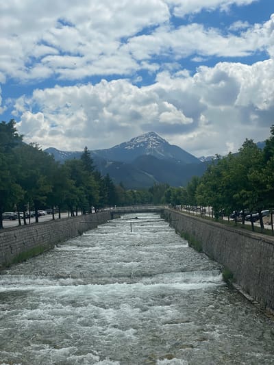 The image depicts a scenic view of the city of Bansko, Bulgaria, featuring a wide, fast-flowing river in the foreground. The riverbanks are lined with stone walls, and on either side, there are rows of green trees. Beyond the trees, a street with parked cars is visible, and further in the distance, a majestic mountain range with snow-capped peaks dominates the skyline. The sky is partly cloudy, with fluffy white clouds against a blue backdrop. A small airplane is visible in the sky. The overall impression is one of natural beauty and urban integration, with the natural landscape playing a prominent role.