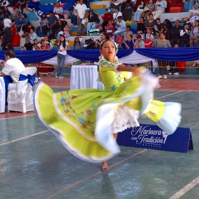 A girl in a green and white dress is captured mid-dance, with the motion causing a blur effect. She has dark hair, fair skin and wears a flowered headpiece. The background includes an audience seated on blue risers with white and blue draped fabric. A sign with the words "Marinera con Tradición REVISTA" is positioned near the dancer. The floor has a green tile-like appearance. Based on the dance and culture shown, it's likely taken in Peru. Though not explicit in the image, Miraflores, Peru has many Marinera festivals.