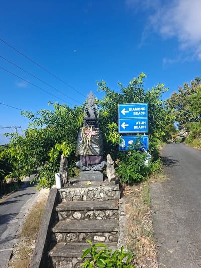 The image depicts a roadside scene in Nusa Penida, Indonesia. In the foreground, a stone staircase leads up to a small shrine adorned with a silver and gold patterned cloth and a fan-like ornament. Behind the shrine, lush green foliage obscures the background. To the right of the shrine, a set of blue directional signs points to "DIAMOND BEACH" and "ATUH BEACH" with white arrows indicating the direction. A paved road runs alongside the foliage and shrine, with a slight incline and dry grass growing along its edge. The sky above is a clear blue with a few wispy white clouds and a crescent moon visible. The overall scene suggests a pathway leading to popular tourist destinations on the island.