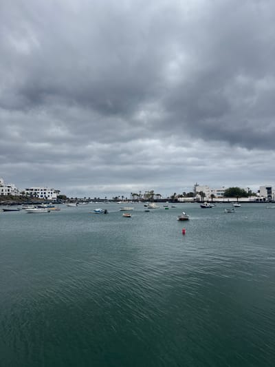 The image shows a harbor in Arrecife, Spain, under a cloudy sky. Several boats of varying sizes are docked along the shoreline and floating in the water. Buildings line the coast, primarily white with some blue accents, characteristic of the region. A red buoy is visible in the water. The water appears calm with slight ripples. There is no visible human activity.