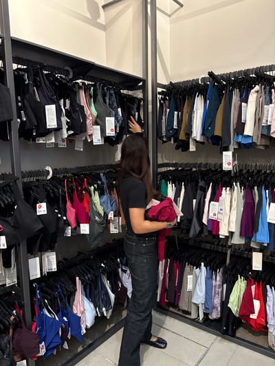 This indoor photograph depicts a retail store environment, likely an athletic wear shop, potentially Lululemon, situated in San Francisco Ocotlán, Mexico. The main subject is a woman with long, dark hair, wearing a dark t-shirt and dark jeans, actively browsing items on a clothing rack. She is seen reaching into a rack, with a folded red garment held in her left arm, and her right hand sifting through other clothing. A silver wristwatch is visible on her left wrist.

The scene is dominated by multiple black metal clothing racks, densely packed with neatly folded athletic wear, including leggings, sports bras, and tops. These garments are displayed in a wide spectrum of colors such as black, various shades of blue, red, green, purple, grey, white, beige, and pink. Each item is attached to a black hanger, and many feature white price tags. Visible text on these tags includes the "lululemon" brand name and logo, along with some red "50% OFF" indicators, suggesting a sale. The store's floor is made of light-colored tiles, and a plain white wall serves as the background, illuminated by bright indoor lighting. The atmosphere suggests a typical daytime shopping experience.