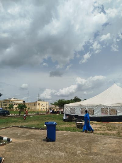 The image shows an outdoor scene in Ukwulu, Nigeria, under a sky with a mix of dark and light clouds. In the foreground, there is a concrete surface, and a blue wheeled trash bin is visible. To the right, a woman in a long blue dress is walking near a large white tent with windows, next to which a generator is placed. Behind the tent, there is grass with people congregated. In the background, there are yellow buildings, trees, street lights, and telecommunication towers. A black car is parked in front of the buildings.