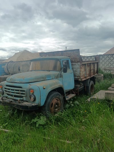 The image depicts an old, dilapidated blue truck parked in a grassy area, presumably in Яльгелево, Russia. The truck has a rusty bed and is surrounded by tall grass and weeds. In the background, there is a pile of sand, a large, cylindrical tank, and a concrete fence. The sky is overcast. No people are visible.