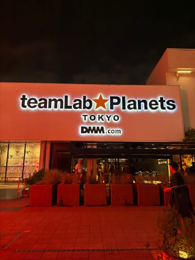The image shows the exterior of teamLab Planets in Toyosu, Koto City, Japan, at night. A large illuminated sign reads "teamLab Planets TOKYO DMM.com". In the foreground, there are large square planters with dried vegetation. The ground is paved with red tiles and illuminated with red light. In the middle ground, there is an entrance to the building, with several people visible inside and entering. Some people are standing near the planters and entrance. To the left of the building, there is a glass-fronted display area with plants and decorative elements inside. The sky is dark.