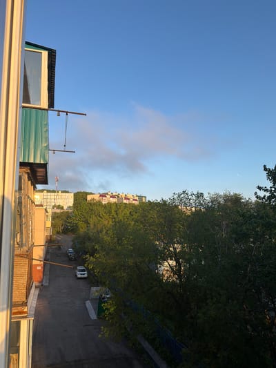 This is a landscape photograph taken from a high vantage point, likely a balcony or rooftop. The image is oriented vertically. The scene depicts a residential area with multiple apartment buildings of varying heights and architectural styles. Trees, lush and green, occupy a significant portion of the right side of the frame, suggesting a natural setting within an urban environment. A paved area, possibly a road or parking lot, is visible beneath the trees, with a few parked cars.
The sky is a clear, bright blue, with some wispy clouds towards the left side of the frame. The lighting suggests it is either early morning or late afternoon, with the sun casting a warm glow on the scene. The overall mood is peaceful and serene.
Notable details include several apartment buildings with a distinct architectural style, some featuring balconies and rows of windows. The trees are dense and full, with sunlight filtering through their leaves. The presence of cars indicates a populated area. There is no visible text in the image. The location context of Petropavlovsk-Kamchatsky, Russia, implies a potentially cooler climate, though the image itself does not provide direct cues to confirm this. The view seems to look out over a neighborhood, capturing a slice of everyday life in this Russian city.