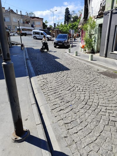 In Sarıyer, Turkey, on a sunlit day, a paved street with cobblestones curves gently to the right. A man wearing a striped shirt and dark pants is seated on a small, dark-colored scooter in the middle of the street, facing away from the viewer. The scooter has two wheels and a flat platform for standing or sitting. To the left of the street, a concrete sidewalk runs parallel, with a grey metal pole, possibly a lamppost, positioned at an angle. Behind the man and scooter, a dark blue SUV is parked on the street, displaying a Turkish license plate that reads "34 HK 595." Further down the street, several white vans are visible, one with "TCDD" written on its side. Buildings line the right side of the street, featuring a light green door and a window with a black frame. Vegetation, including a potted plant, grows alongside the buildings. The sky is blue with scattered white clouds.